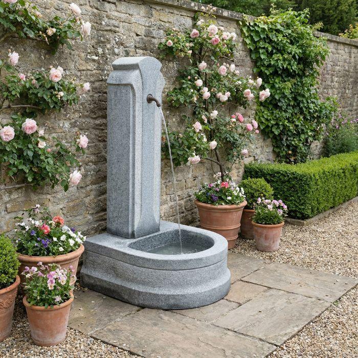 Fontaine en granit dans un jardin avec roses grimpantes et pots en terre cuite